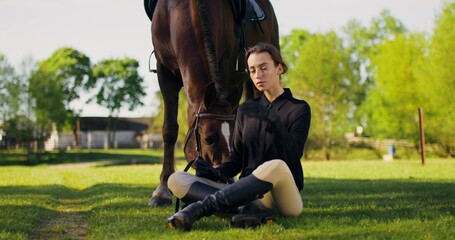 A stylish woman is sitting on the grass next to a horse. She poses looking at the camera