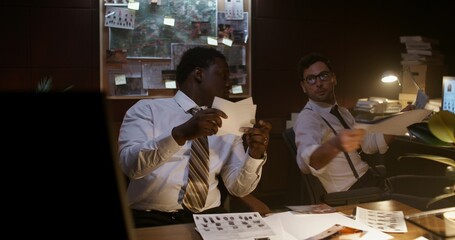 Two detectives are concentrating on working in the office. They study crime scene photos and talk while sitting at their desks.