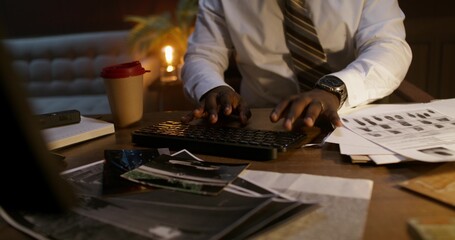 The detective uses a computer while sitting at his desk in the office. Photographs and evidence on the table in front of him
