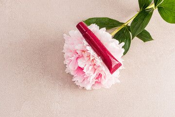 An unnamed tube of cosmetics lies on a large gorgeous pink peony flower. Beige stone background. A...