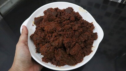 A hand holding a white plate filled with rendang, dark brown, savory cuisine, traditional food from Indonesia. 