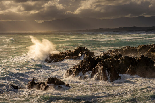 Powerful waves crash against rugged rocks at De Kelders, Western Cape, South Africa, under dramatic skies. The wild ocean and scenic coastline create a breathtaking seascape at sunset.