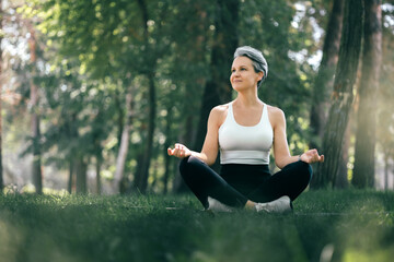 Fototapeta premium Smiling Mature Woman Meditating Outdoors in Sukhasana Yoga Pose Surrounded by Nature