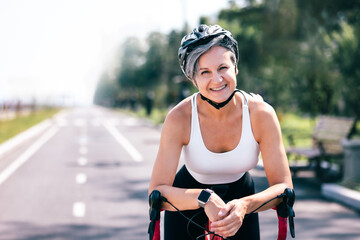 Happy senior woman cyclist in helmet leaning on bicycle on sunny road with confident smile