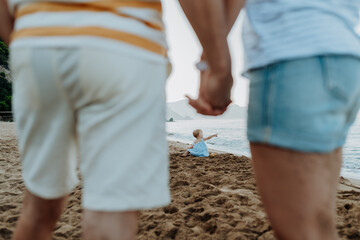 Parents looking at little toddler girl sitting on beach during beach holiday.