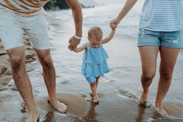 Parents holding hands with little toddler girl, walking on beach during beach holiday.