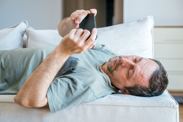 Lazy couch potato guy using mobile phone while laying on living room sofa