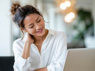Young Asian businesswoman suffering neck pain from working on a laptop at her office desk. The woman, wearing a white shirt, is experiencing upper back or shoulder discomfort. The