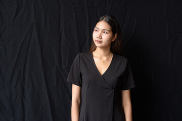 Fototapeta premium Asian woman in black dress posing against black curtain in natural light