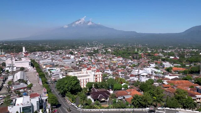 A cityscape of Salatiga with urban buildings in the foreground, framed by the majestic Mount Merbabu rising peacefully in the background under a clear sky.
