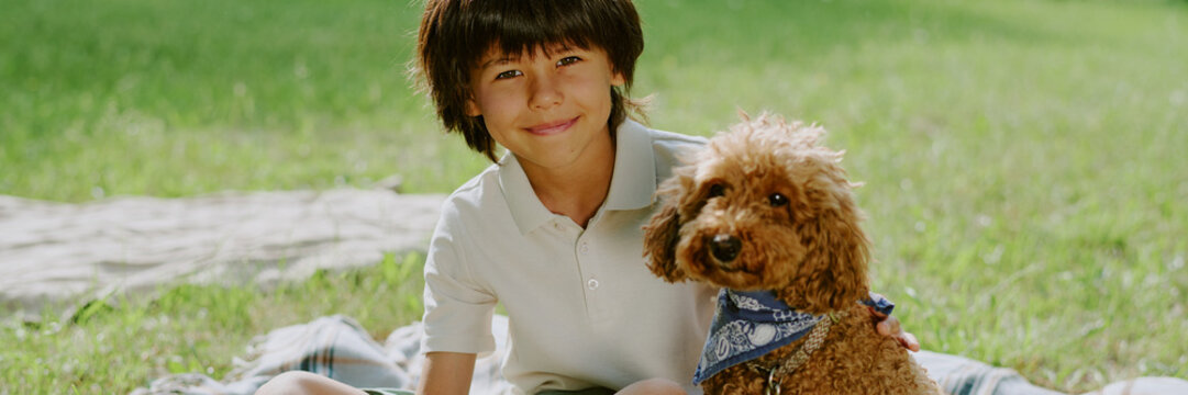 Portrait of biracial boy sitting on grass in park smiling at camera with curly brown dog wearing bandana, child gently holding pet outdoors during sunny day - Powered by Adobe