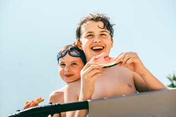 Two cheerful boys eating watermelon together and enjoying a sunny summer day in the pool