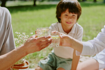 Biracial child sitting on grass in park smiling while two adults clinking glasses of water in foreground, hands of young adult man and woman visible, outdoor family gathering