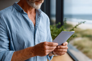 Senior man reading a letter by the window overlooking the ocean
