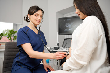 Female doctor listening to pregnant woman's belly with stethoscope during prenatal checkup. Medical...