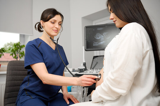 Female doctor listening to pregnant woman's belly with stethoscope during prenatal checkup. Medical care and fetal health monitoring during pregnancy.