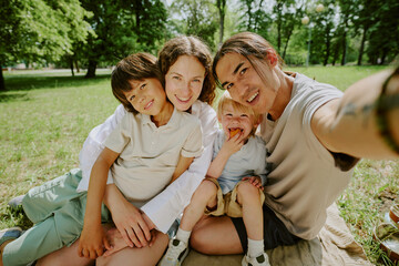 Portrait of Caucasian woman, Asian man and their biracial children sitting closely together on grass in park smiling at camera, parents embracing children outdoors