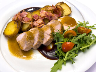 Pork tenderloin with roasted potatoes, bacon and plum sauce, served with fresh arugula and cherry tomatoes. Tasty meat dish on white plate, top view.
