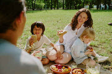 Caucasian woman sitting on grass with two children and young adult man having picnic, smiling and eating fruit, biracial kids enjoying outdoor family time in park on sunny day