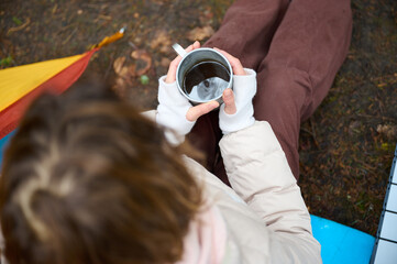 Hiker woman drinking coffee outdoor near the camping tent. A female traveler in the forest in early...