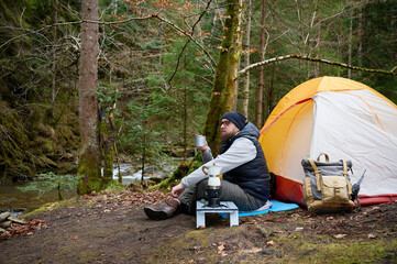 Man hiker sitting near camping tent and holding a mug with hot coffee. Adventure, travel, tourism...
