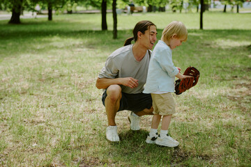 Asian young adult man kneeling beside his child boy teaching him how to use baseball glove in park,...
