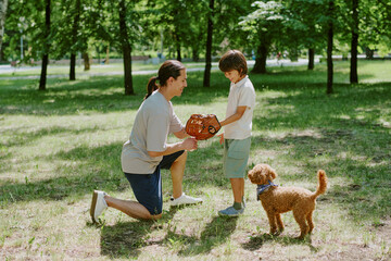 Asian young adult father kneeling and teaching son how to use baseball glove in park, while curly...