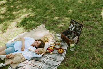 Caucasian middle aged woman and child lying on picnic blanket in park relaxing together near basket...