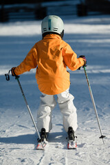Child in bright orange jacket prepares to ski down snowy slope during a sunny winter afternoon,...