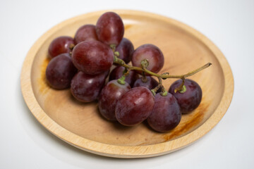 A wooden plate filled with fresh, juicy red grapes placed on a white background.