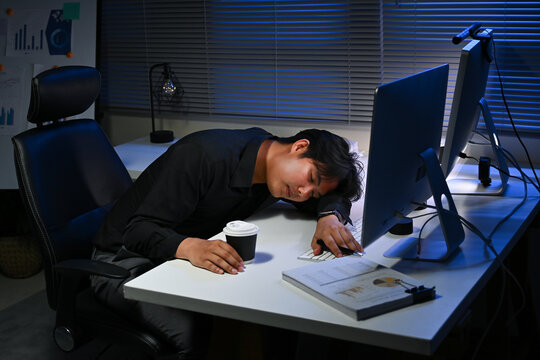 A man is sleeping on a desk with a computer monitor and a cup of coffee. The scene suggests that the man is working late and is exhausted - Powered by Adobe