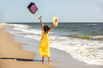 Young happy woman in a yellow long dress with hands in the air runs on the seaside in summer. The...