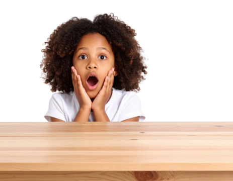 A young girl with curly hair shows a shocked expression, holding her face with both hands while sitting behind a wooden table against a black background.
