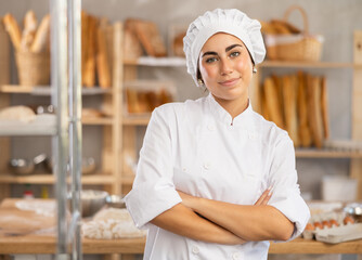 Cheerful young female baker stands in bake house, ready to greet customers