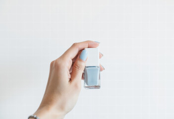 A woman's hand with a fresh, light blue manicure holds a matching bottle of nail polish against a clean white background, showcasing a chic, minimalist style.
