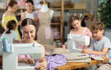 Teen girl child sews during practical lesson at school of young fashion designers and tailors. Classmates are doing preparatory work together in background.