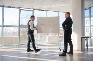 Two Caucasian men discussing work at a white board. 