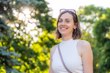Portrait of cheerful woman smiling in park with sunlight streaming through trees, embodying...