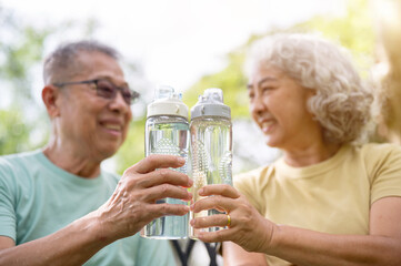 Close up of old man and woman holding and clinking their water bottles together after exercise in park.