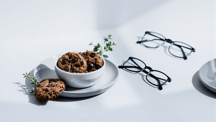 Close-up of a bowl of chocolate chip cookies with a pair of eyeglasses on a white surface, highlighting a cozy and casual setting with minimalistic background and natural lighting