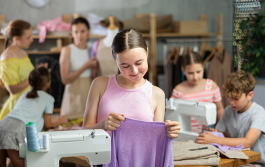 Girl child with cloth in hands examines product, school of young fashion designers. Students in...