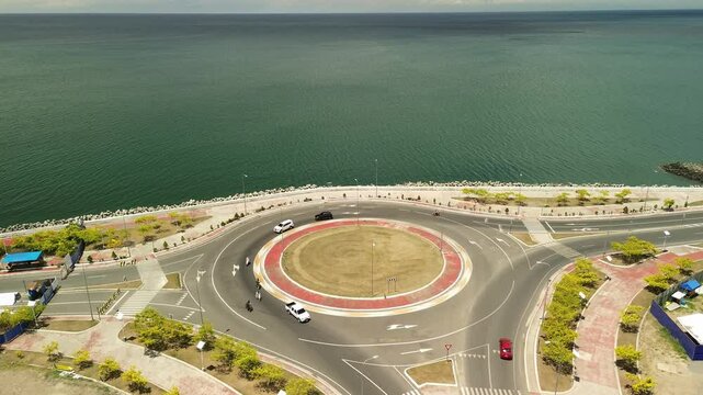 Slow drone ascent showcases a sunlit traffic circle on Davao City&rsquo;s seafront boulevard, vehicles tracing the ring road against shimmering Mindanao Sea.