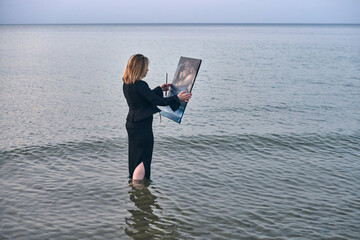 Young woman stands knee-deep in calm ocean water, concentrating on her canvas as she captures...