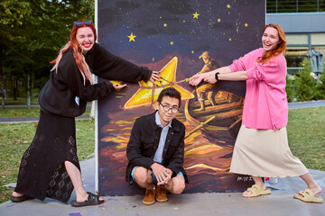 Two young female artists and young male pose playfully with outdoor painting in park. Vibrant...