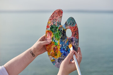 Young female holds colorful artist palette against tranquil seascape, symbolizing creativity and...
