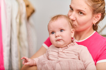 Young mother with light brown hair, holding baby girl. Woman wears pink top, baby in light dress. Warm emotions, indoor environment, bright lighting