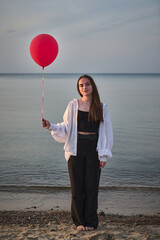 Young woman stands on quiet beach holding red balloon. Serene seascape and soft evening light...