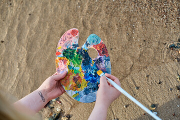 Young adult holds vibrant paint palette, blending colors with brush on sandy beach, embodying...