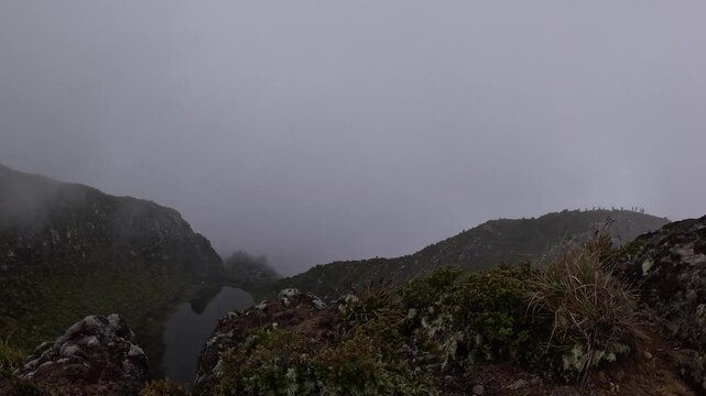 First-person ridge walk passes a lichen-stained boulder toward Mount Apo&rsquo;s fog-veiled second summit, narrow dirt path threading rugged slopes under a muted grey sky.
