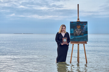 Young woman stands beside easel on serene beach, with calm sea and overcast skies creating...